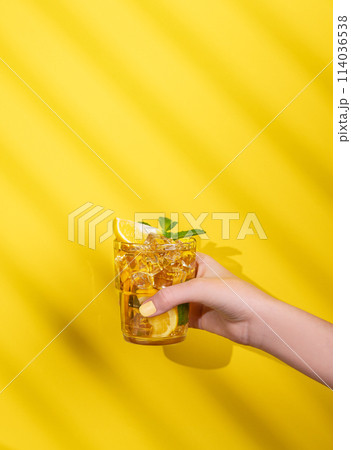 A hand holds a glass of iced tea with lemon and mint on a yellow background with shadow. A hand holds a glass of iced tea with lemon and mint on a yellow background with shadow. 114036538