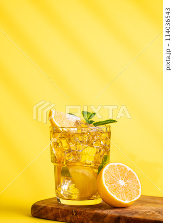 A glass of iced tea with lemon and mint on a wooden board on a yellow background with a shadow. A glass of iced tea with lemon and mint on a wooden board on a yellow background with a shadow. 114036543