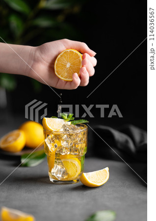 A hand squeezes juice from a lemon into a glass with lemonade with ice and mint on a dark background 114036567