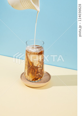 Iced coffee in a tall glass with a milk jug pouring milk on a blue and yellow background with shadow 114036628