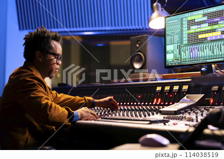African american sound engineer works on mixing console with sliders and knobs, looking at digital software to record and process tunes. Audio technician creating new music, control room board. African american sound engineer works on mixing console with sliders and knobs, looking at digital software to record and process tunes. Audio technician creating new music, control room board. 114038519