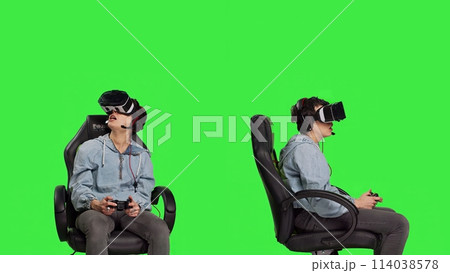 Woman playing video games on console with controller and vr headset, sitting against greenscreen backdrop. Gamer having fun with online tournament, virtual reality interactive cyberspace. Camera B. 114038578