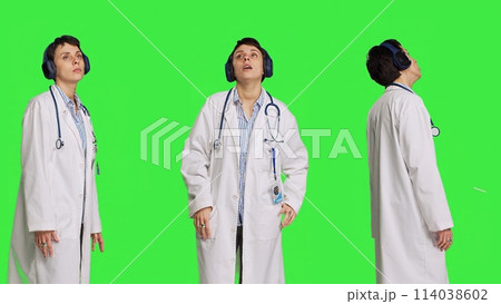 Joyful physician dancing around in the studio with music on headset, having fun with modern cool songs against greenscreen backdrop. Medic woman enjoying audio tunes and singing. Camera B. 114038602