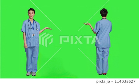 Medical assistant pointing at something aside and doing web commercial, creating advertisement against greenscreen backdrop. Young nurse with scrubs showing a thing to left or right sides. Camera A. 114038627