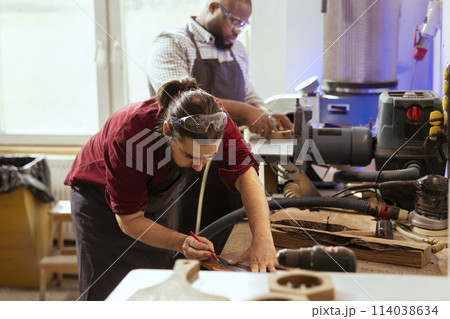 Woodworker using ruler to take measurements of wood plank with african american coworker. Creative person drawing with pencil on wooden board to determine where to make cut, helped by apprentice Woodworker using ruler to take measurements of wood plank with african american coworker. Creative person drawing with pencil on wooden board to determine where to make cut, helped by apprentice 114038634