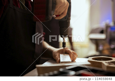 Carpenter drilling hole into timber block, doing furniture assembling in studio, close up. Woodworking expert in joinery inserting nail into piece of wood using electric power drill tool 114038648