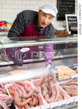 Confident male butcher holding large beef sausage in meat section of supermarket Confident male butcher holding large beef sausage in meat section of supermarket 114042797