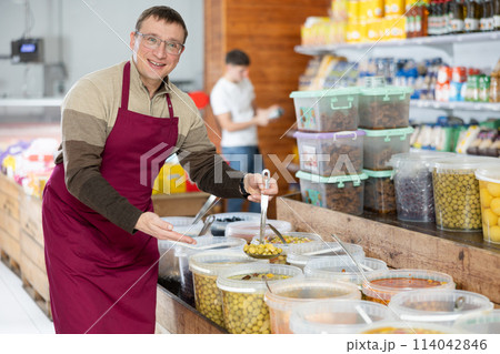 Man in supermarket, scooping marinated olives from bucket with ladle 114042846