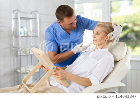 Senior woman looking at mirror while cosmetologist explaining facial procedure Senior woman looking at mirror while cosmetologist explaining facial procedure 114042847
