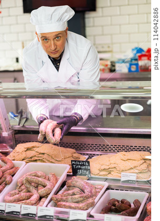 Confident male butcher holding large beef sausage in meat section of supermarket 114042886