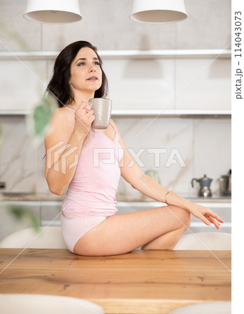 Woman in white lady singlet lingerie and underpants is sitting on kitchen table Woman in white lady singlet lingerie and underpants is sitting on kitchen table 114043073