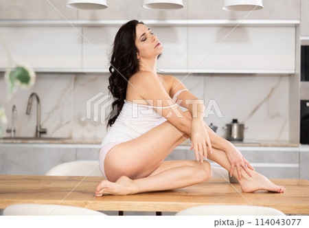 Portrait of happy brunette young woman wearing underwear playfully posing on table at white kitchen Portrait of happy brunette young woman wearing underwear playfully posing on table at white kitchen 114043077