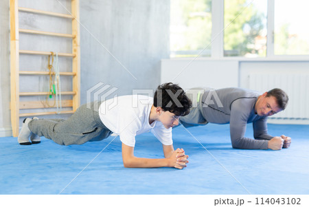 Father and son train abdominal muscles in gym - perform plank exercise 114043102