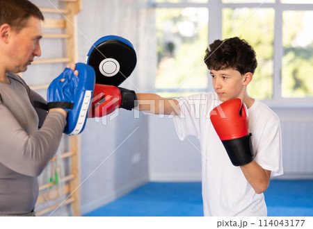 Teenage boy training boxing kicks on punch mitts held by instructor 114043177