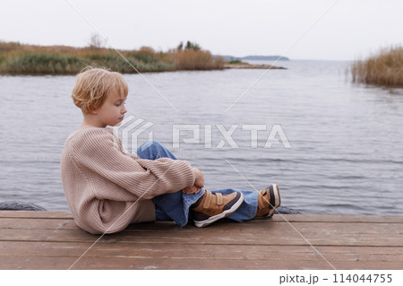 A young girl sits on a dock by a body of water, wearing a sweater and jeans 114044755