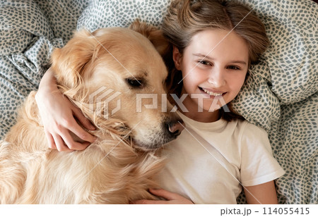 Happy Little Girl Hugging Golden Retriever Dog And Smiling Lying On A Bed 114055415