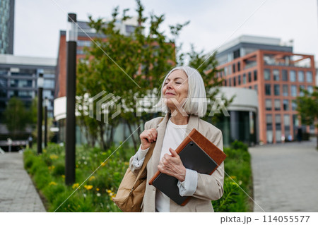 Mature businesswoman going home from work, enjoying beautiful weather and free time. Beautiful older woman with gray hair walking down city street. 114055577