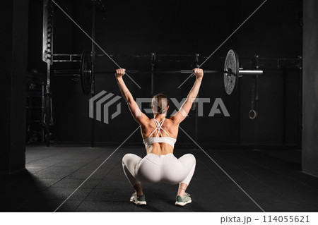 Rear view of woman performing overhead squat pressing barbell up, standing up. Routine workout for physical and mental health. 114055621