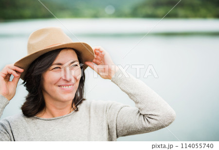 Portrait of beautiful mature woman with hat, standing by lake. Portrait of beautiful mature woman with hat, standing by lake. 114055740