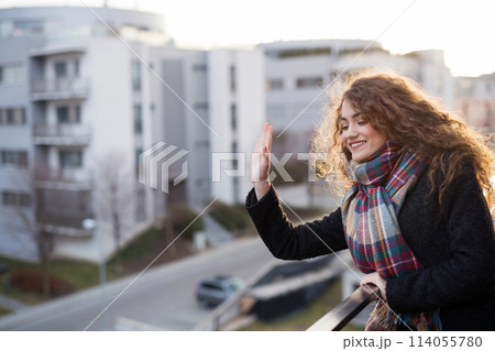 Beautiful woman with curly hair standing on terrace, waving someone down at street. Morning sun during cold spring day. Beautiful woman with curly hair standing on terrace, waving someone down at street. Morning sun during cold spring day. 114055780