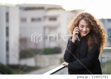 Beautiful woman with curly hair standing on terrace, making phone call outdoors during cold spring day. Morning sun, hair blowing in wind. Beautiful woman with curly hair standing on terrace, making phone call outdoors during cold spring day. Morning sun, hair blowing in wind. 114055781