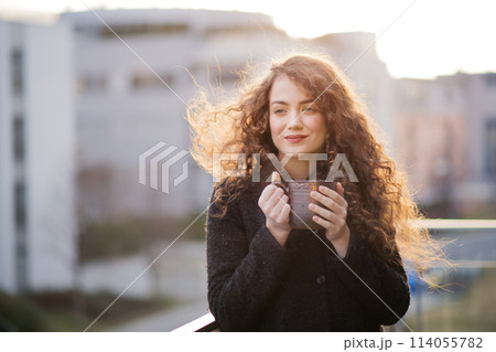 Beautiful woman with curly hair standing on terrace, drinking coffee during cold spring day. Morning sun, hair blowing in wind. 114055782