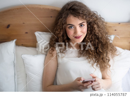 Beautiful woman with curly hair sitting on bed, scolling on smartphone. Beautiful woman with curly hair sitting on bed, scolling on smartphone. 114055787