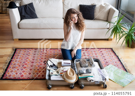 Beautiful woman planning summer vacation abroad, sitting on floor, packing suitcase. Solo traveler making phone call. 114055791