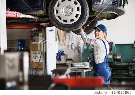 Female auto mechanic elevating car on car lift, working underneath. Beautiful woman working in a garage, wearing blue coveralls. Female auto mechanic elevating car on car lift, working underneath. Beautiful woman working in a garage, wearing blue coveralls. 114055863