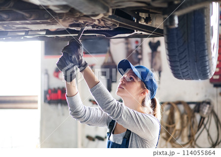 Female auto mechanic elevating car on car lift, working underneath. Beautiful woman working in a garage, wearing blue coveralls. 114055864