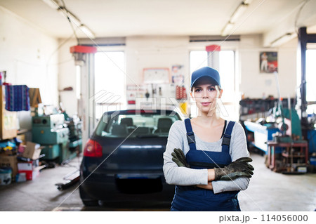 Female auto mechanic repairing, maintaining car. Beautiful woman standing in a garage, wearing blue coveralls. Female auto mechanic repairing, maintaining car. Beautiful woman standing in a garage, wearing blue coveralls. 114056000