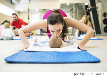 Mother smiling at baby while performing push-ups, kissing infant. Group exercise class, fitness or pilates for parents. Moms staying active while boding with babies. Mother smiling at baby while performing push-ups, kissing infant. Group exercise class, fitness or pilates for parents. Moms staying active while boding with babies. 114056054