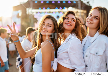 Group of happy girls at summer beach music fest, smiling, dancing in sun, flashing peace sign. Friends enjoy live concert, sunset light in background. Casual trendy outfits, fun, youth culture vibe. 114056260