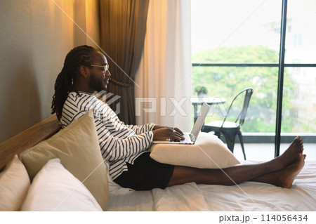 Young African American man using laptop computer on bed , working at his hotel room 114056434