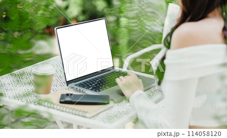 Over-the-shoulder view of woman using laptop with blank screen on outdoor table, with coffee and smartphone. 114058120