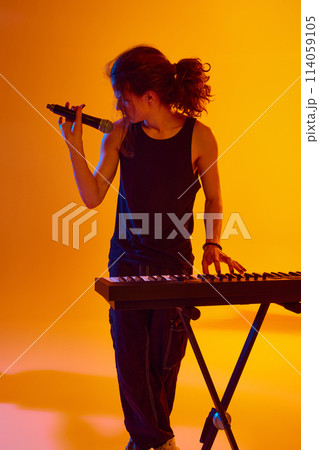 Young man making soulful performance, playing piano and singing against yellow background in neon light 114059105