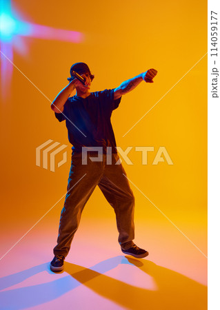 Young emotional guy in casual clothes standing on stage, reading rap against yellow background in neon light Young emotional guy in casual clothes standing on stage, reading rap against yellow background in neon light 114059177