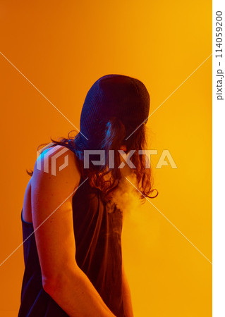 Punk rock. Young man with long hair, wearing black balaclava, smoking on stage against yellow background in neon light Punk rock. Young man with long hair, wearing black balaclava, smoking on stage against yellow background in neon light 114059200