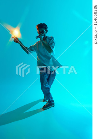 With determined expression, young musician delivers powerful rap performance against blue, cyan background in neon light 114059286