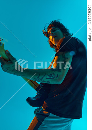 Young man, bathed in blue light, playing electric guitar, making energetic rock and roll performance against blue, cyan background in neon light Young man, bathed in blue light, playing electric guitar, making energetic rock and roll performance against blue, cyan background in neon light 114059304