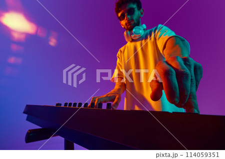 Young man, lost in the rhythm, mixes tracks on a DJ mixer, fueling energy of crowd against gradient pink purple background in neon light Young man, lost in the rhythm, mixes tracks on a DJ mixer, fueling energy of crowd against gradient pink purple background in neon light 114059351