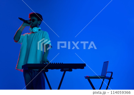 Dimly lit club scene with young DJ energizing the crowd with infectious music against blue background in neon light 114059366