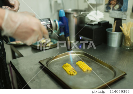 Cook grills sweet corn with handy gas burner in kitchen Cook grills sweet corn with handy gas burner in kitchen 114059393