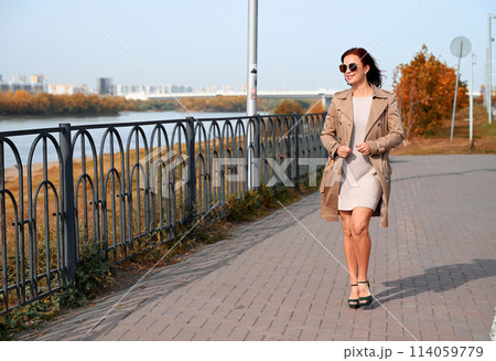 a confident young attractive woman in a coat and high-heeled shoes walks through the streets of the city, the woman smiles and enjoys life 114059779