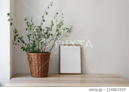 Spring, Easter still life. Elegant Scandinavian living room, home office. Empty vertical wooden picture frame mockup on desk, table. Wicker willow basket with green birch tree branches. Front view. 114061507