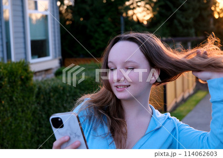 girl at sunset straightens hair and looks at screen of phone getting ready for photo shoot take selfie reflection bite little lip smile wait for phone call from friends backlight of sun on hair girl at sunset straightens hair and looks at screen of phone getting ready for photo shoot take selfie reflection bite little lip smile wait for phone call from friends backlight of sun on hair 114062603