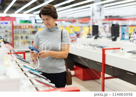 Elderly female customer looking for a deal on phones in big electronics store 114063056