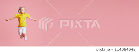 Banner. Boy jumping of joy while listening music in headphones against pink studio background with negative space to insert text. 114064048