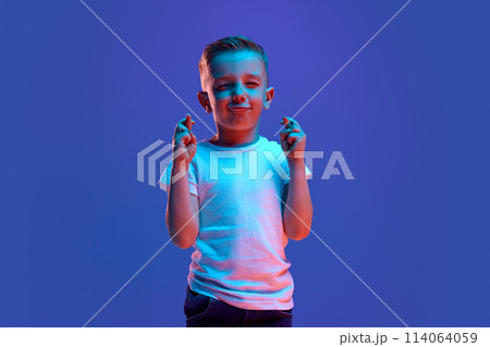 Good luck at exam. Cute child, boy crossed fingers and closed eyes to get lucky in mixed neon light against blue background. 114064059