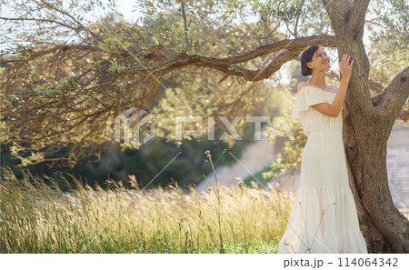 Beautiful Asian young woman in white dress outdoor near olive tree. embracing fresh air and engaging in outdoor activities. Friluftsliv concept means spending as much time outdoors as possible Beautiful Asian young woman in white dress outdoor near olive tree. embracing fresh air and engaging in outdoor activities. Friluftsliv concept means spending as much time outdoors as possible 114064342